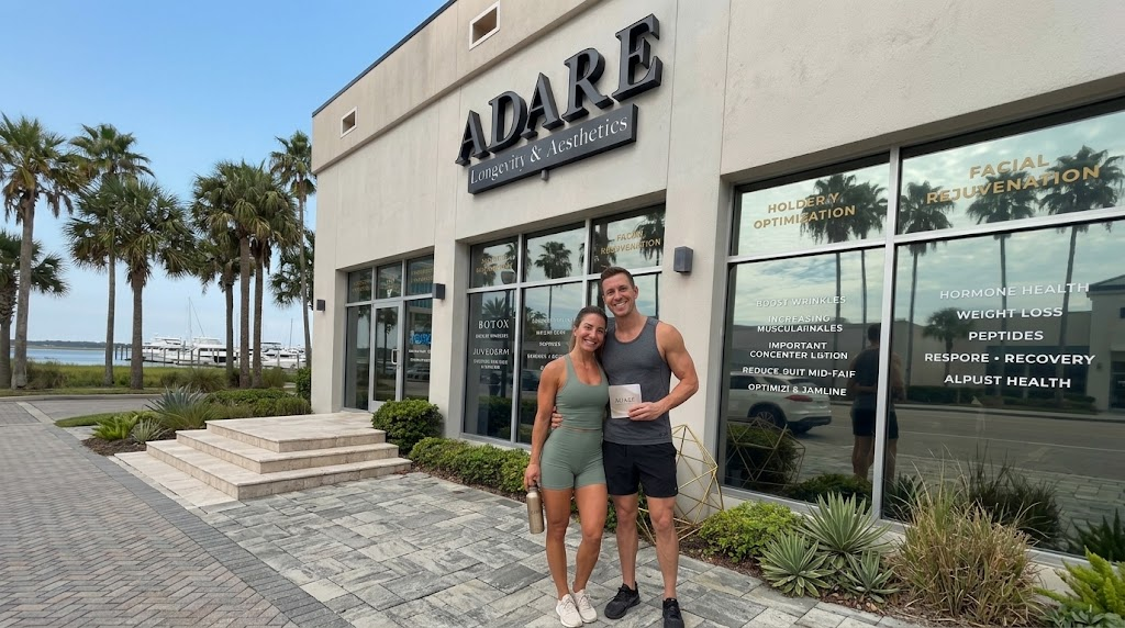 A fit couple with athletic physiques poses with broad smiles and hugs in front of the ADARE Longevity & Aesthetics studio facade in a sunny Florida setting. The woman wears sage green athletic wear and holds a branded water bottle, while the man wears a grey tank and black shorts and holds an information brochure.