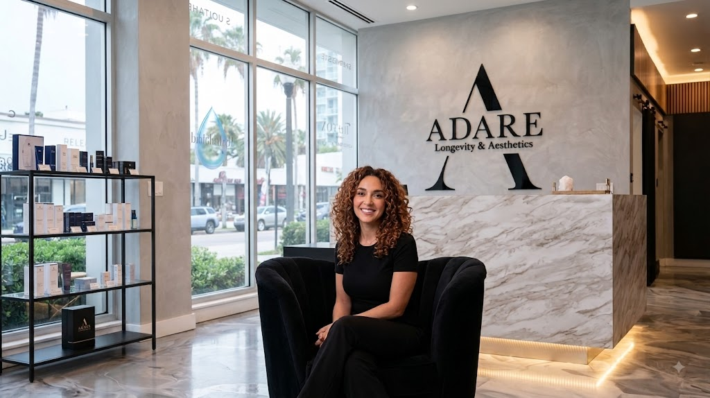A smiling woman with curly hair sitting in a black velvet chair in a luxurious clinic reception area, with a large marble desk and the 'ADARE Longevity & Aesthetics' logo in the background.
