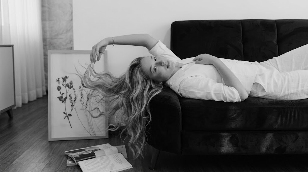 A thoughtful black and white photograph of a woman with long, flowing hair resting her head on a dark velvet sofa and gazing directly at the camera. Her hand is raised in a pensive gesture, and she wears a white button-up shirt and vest. An open book and a framed botanical print are visible on the wooden floor nearby.