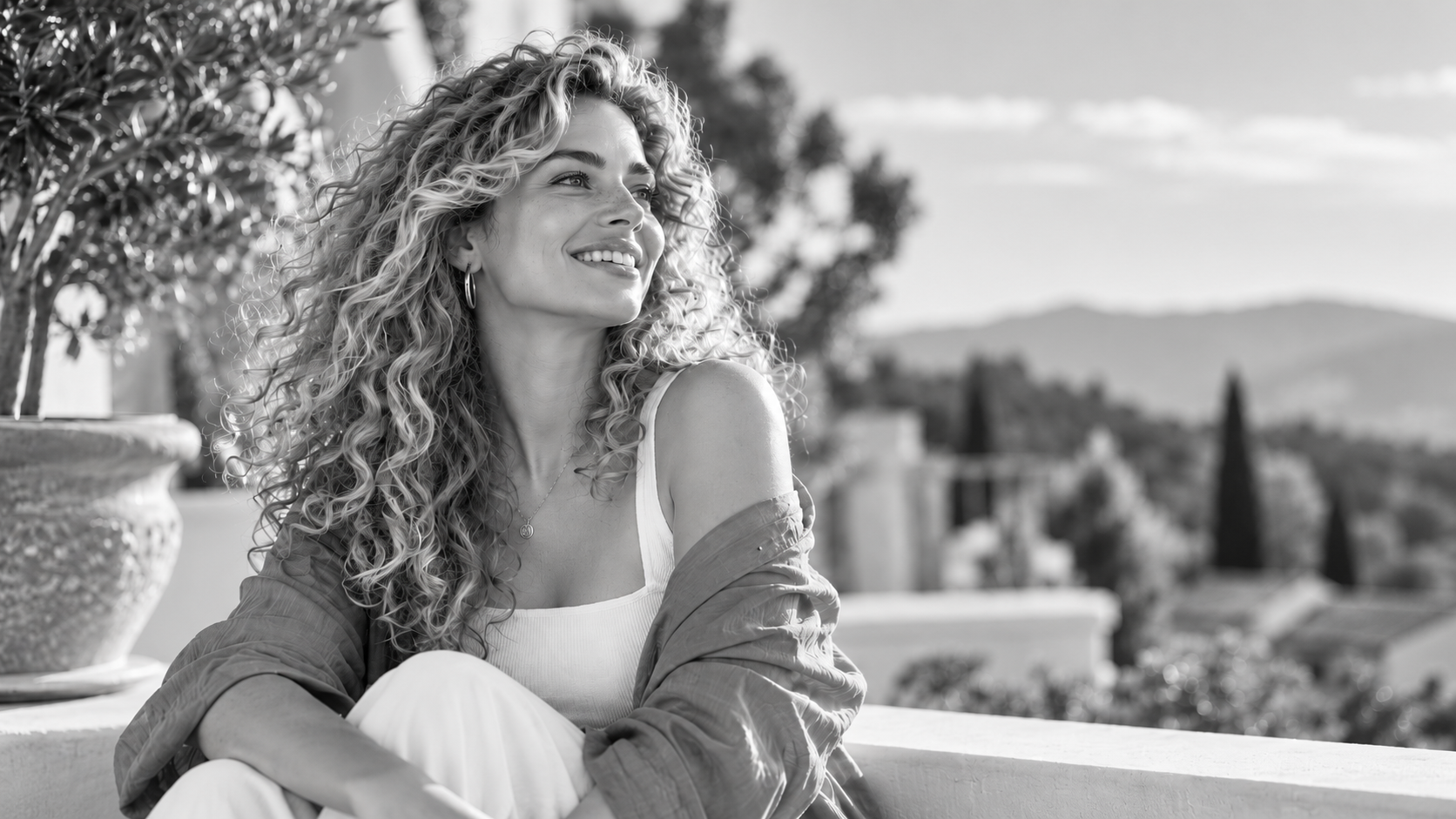 Smiling blonde woman with long curly hair sitting on a sunlit terrace in a high-quality grayscale portrait.