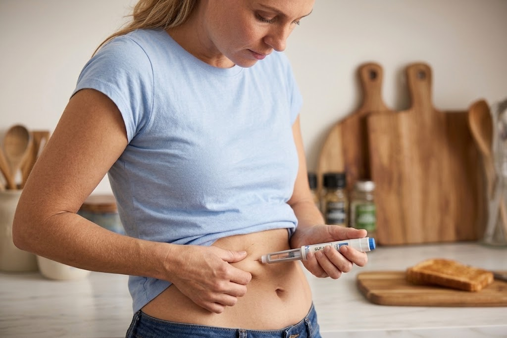 A woman in a blue t-shirt and jeans pinches her abdominal skin and holds a GLP-1 injection pen, about to administer the medication in a kitchen setting.