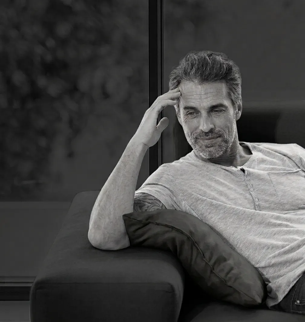 A high-contrast black and white photograph of a diverse mature man with textured grey hair and a full beard, reclining on a dark sofa. He is wearing a marled t-shirt with rolled sleeves, revealing intricate patterned tattoos. One hand rests thoughtfully near his temple, and he is gazing downward and to the right with a soft, pensive smile.