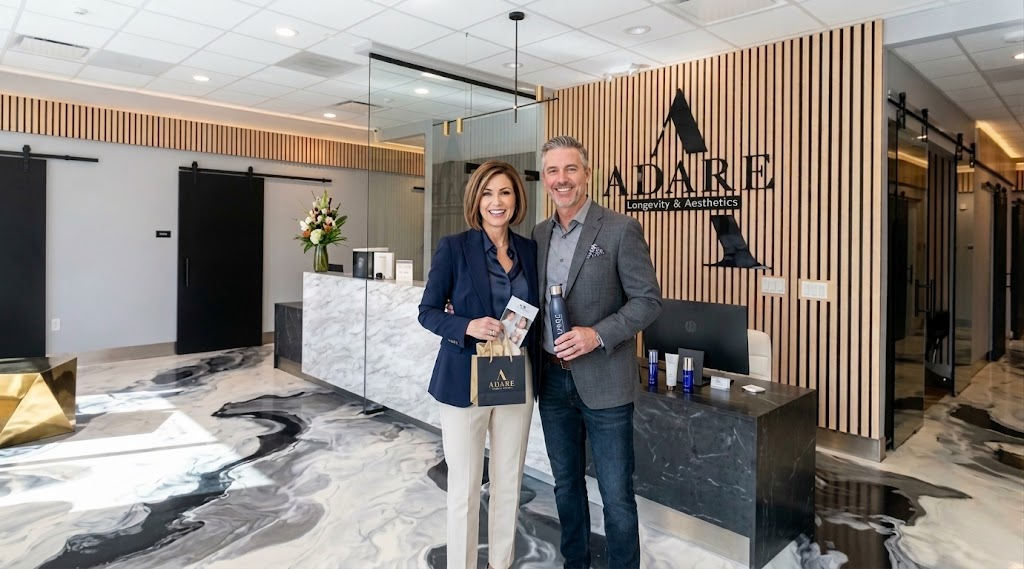 A man and a woman standing in the modern reception area of ADARE Longevity & Aesthetics. The woman holds a branded paper shopping bag, and the man holds a branded water bottle next to a reception desk with a wooden slatted wall background.