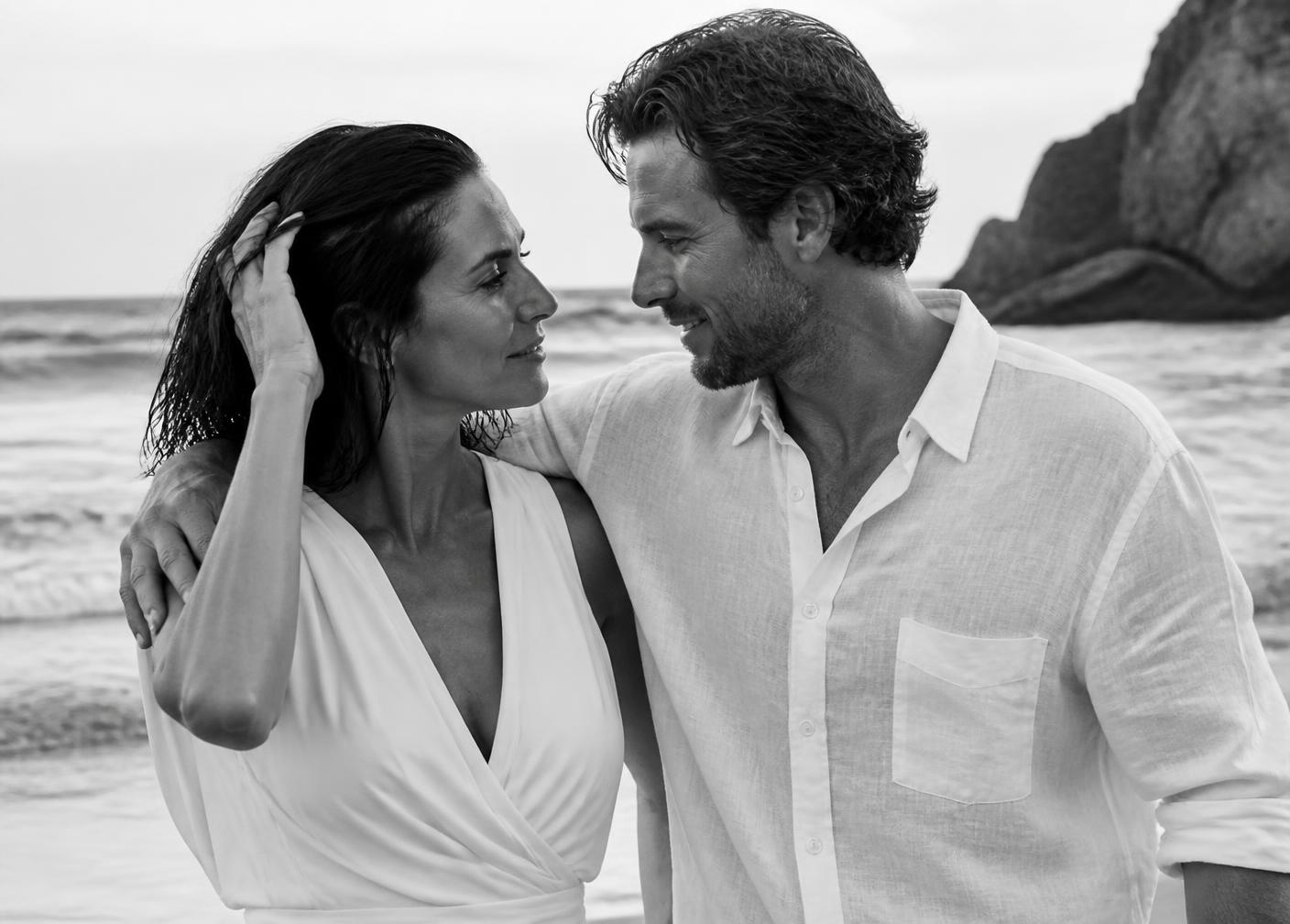 A black and white portrait of a couple in their early 40s gazing into each other's eyes on a beach. The woman wears a sleeveless white dress and rests her hand near the man's hair, while the man wears a white button-down linen shirt with his arm around her shoulder.