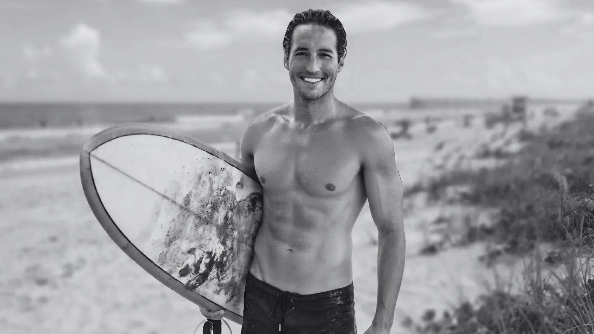 A medium shot photograph of a smiling, shirtless muscular man with wet, slicked-back hair, holding a worn, sand-covered surfboard under his left arm. He is standing on a sun-drenched beach with sand dunes and sea oats in the foreground, and a blurred ocean, waves, a distant pier, and beachgoers in the background under a partly cloudy sky. The man is looking directly at the camera.