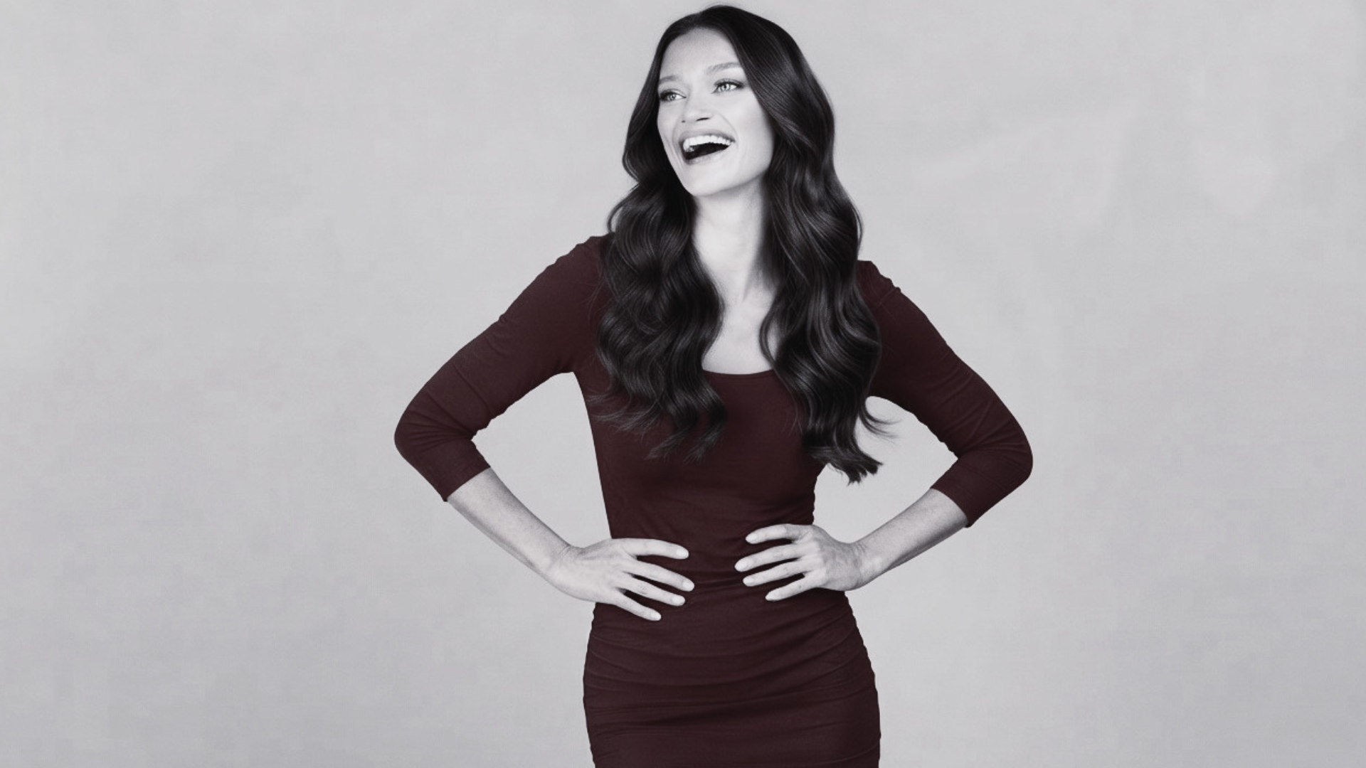 A medium shot photograph of a joyful woman with long, wavy brown hair, wearing a form-fitting, ruched maroon or deep red dress with three-quarter sleeves. She is standing with her hands on her hips and looking off-camera to her left with a wide, open-mouthed smile. The background is a solid, neutral, light beige or tan studio backdrop. The lighting is soft and even.