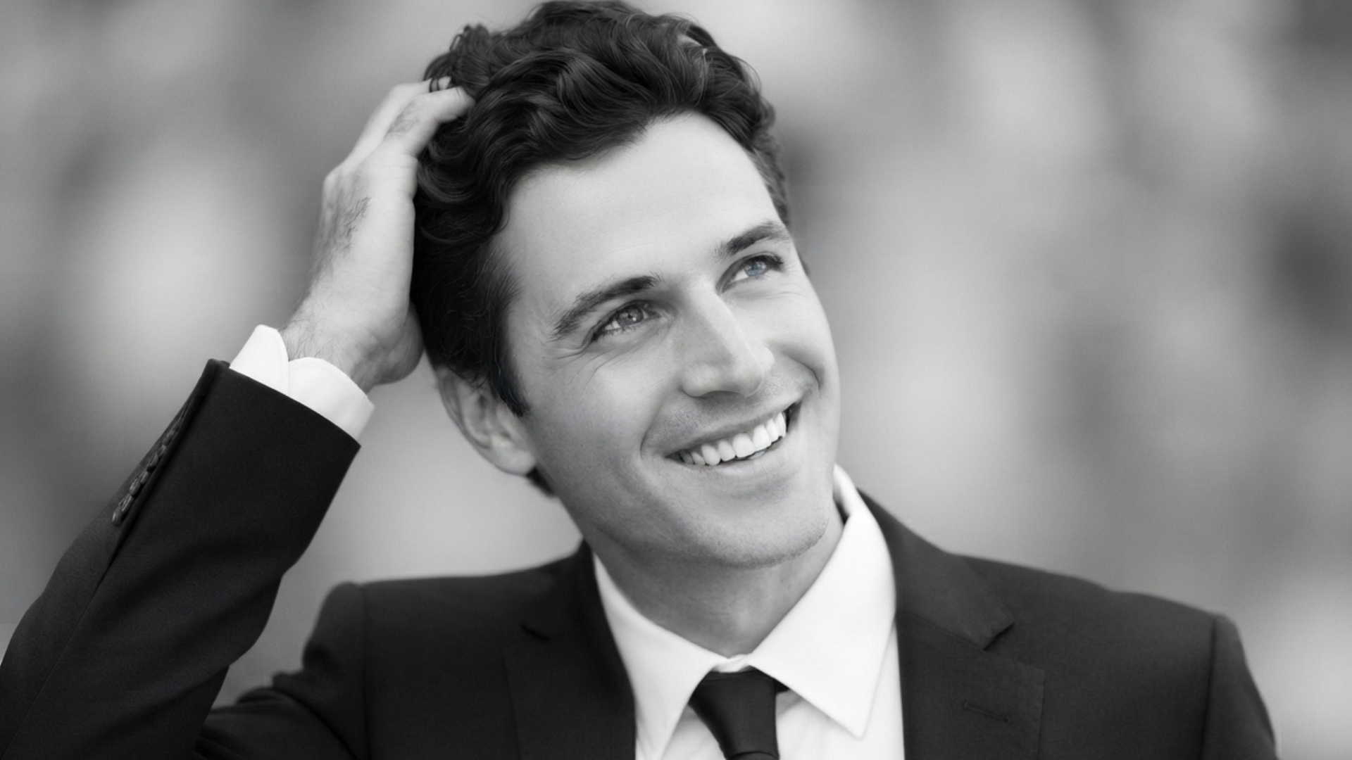 A black and white photograph capturing a smiling man in a suit looking upwards and to the side with his hand in his hair.