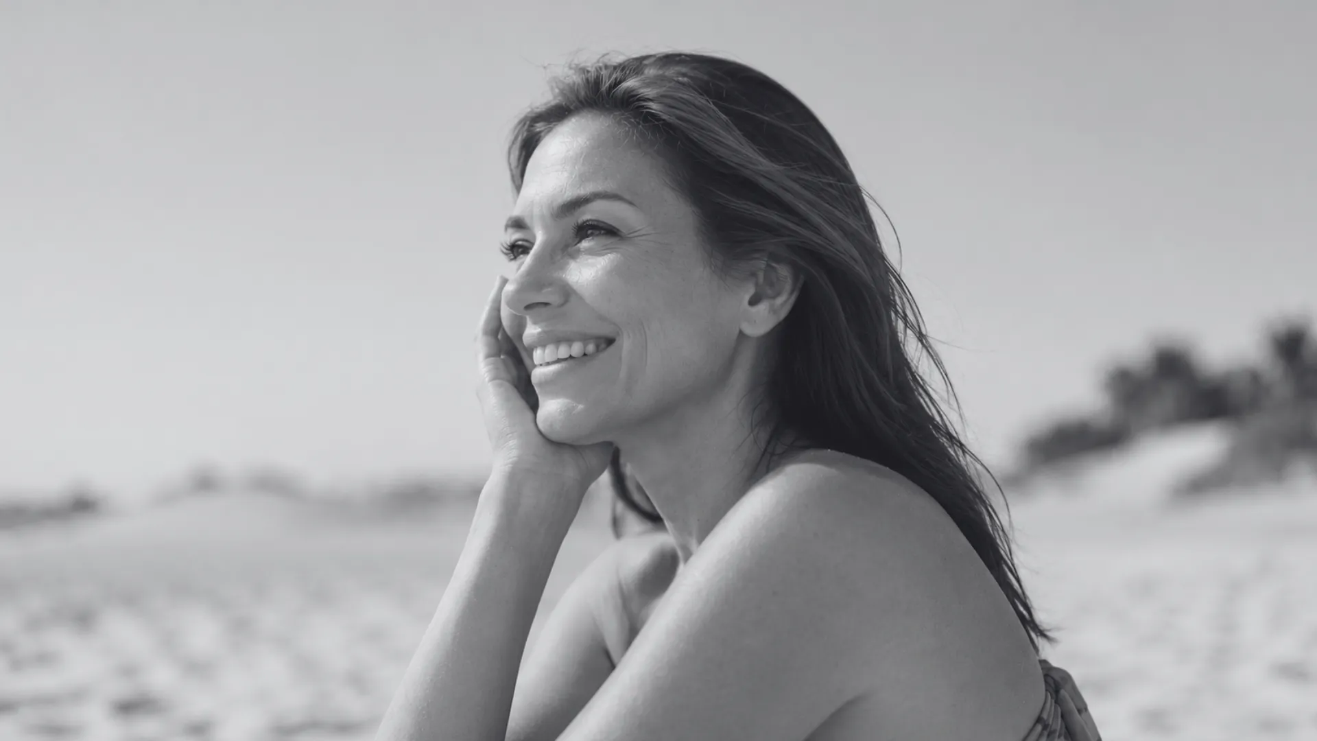 A monochrome landscape photograph of a smiling woman in her 40s resting her chin in her hand, looking off to the side with a joyful expression. She is outdoors at a beach setting with a softly blurred background.